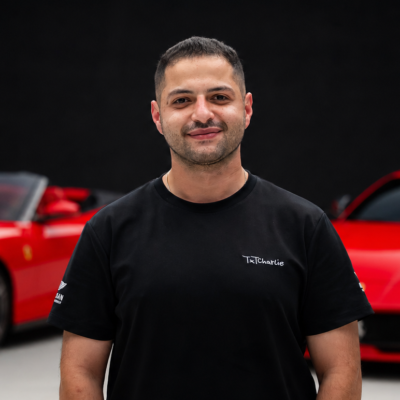Portrait of a smiling man in a black T-shirt with a white 'TexCharlie' logo, standing in front of red cars indoors.