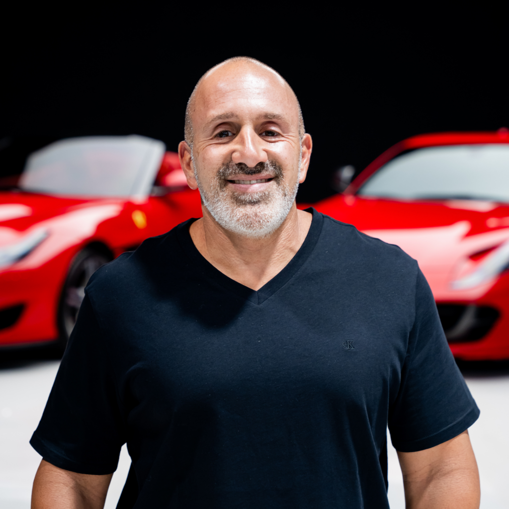 1png | Txtcharlie Smiling man with a shaved head and gray beard in a black V-neck shirt, standing in front of blurred red cars.