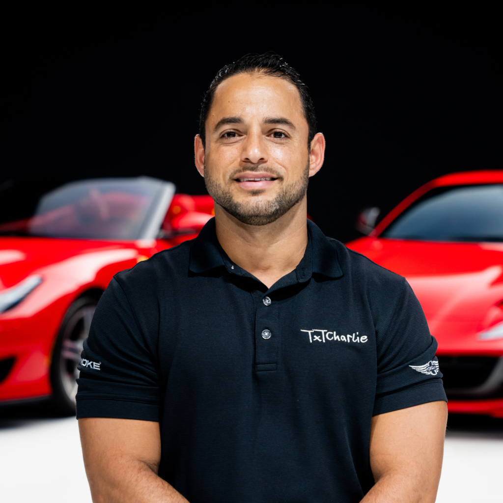 11png | Txtcharlie Close-up portrait of a man in a black polo shirt with a small logo, standing in front of red cars against a dark background.