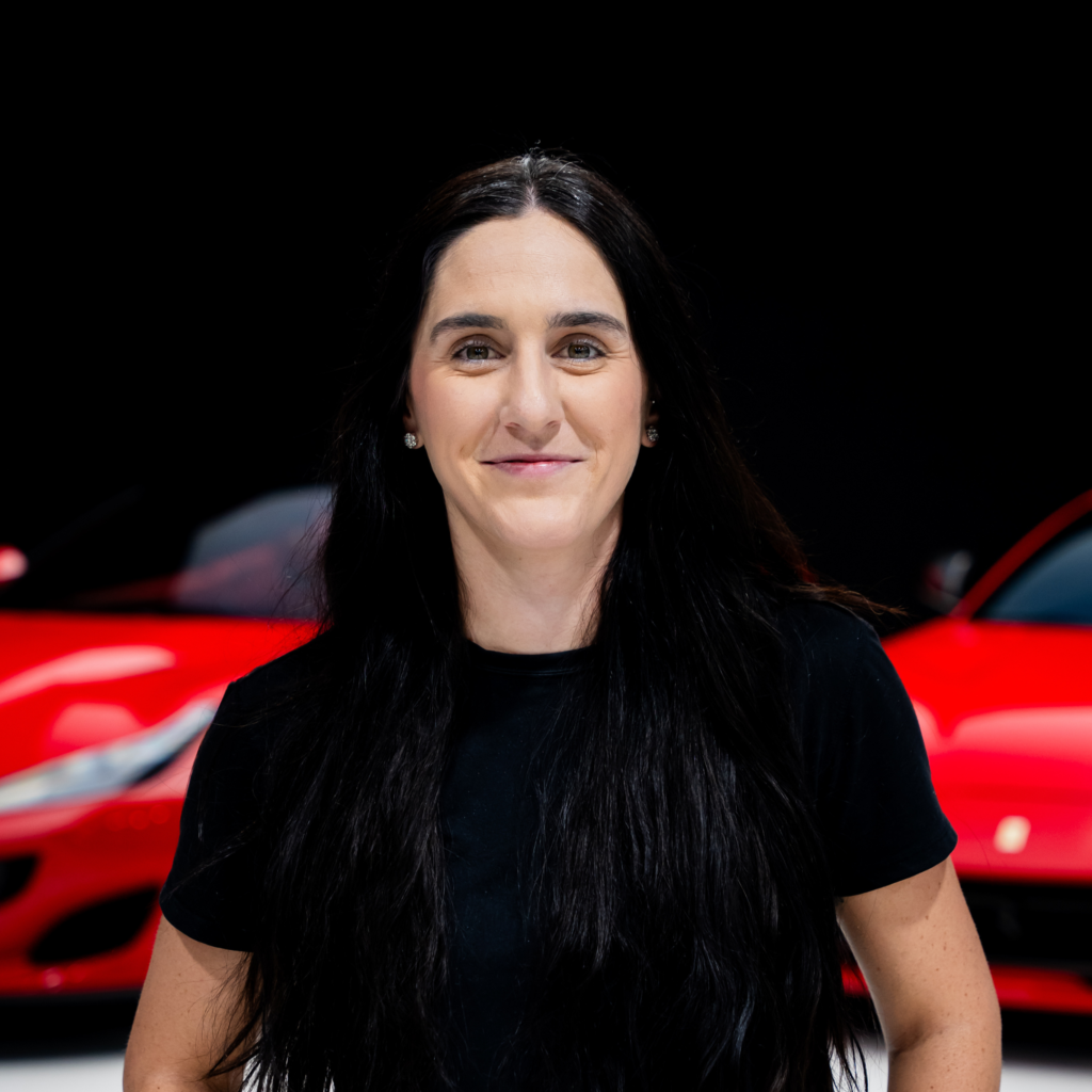 142png | Txtcharlie Close-up of a smiling woman with dark hair and a black shirt, against a black background with a red car blurred in the foreground.