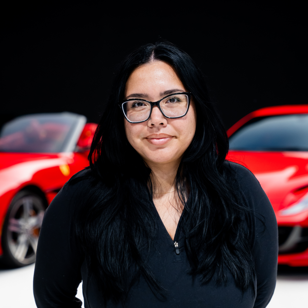 16png | Txtcharlie Close-up of a woman with black hair and glasses, wearing a black top, standing in front of a red car in the background.