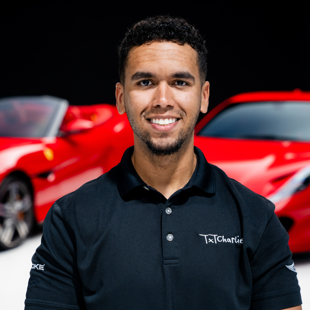 2png | Txtcharlie Smiling man in a black polo standing in front of a red car with a dark background.