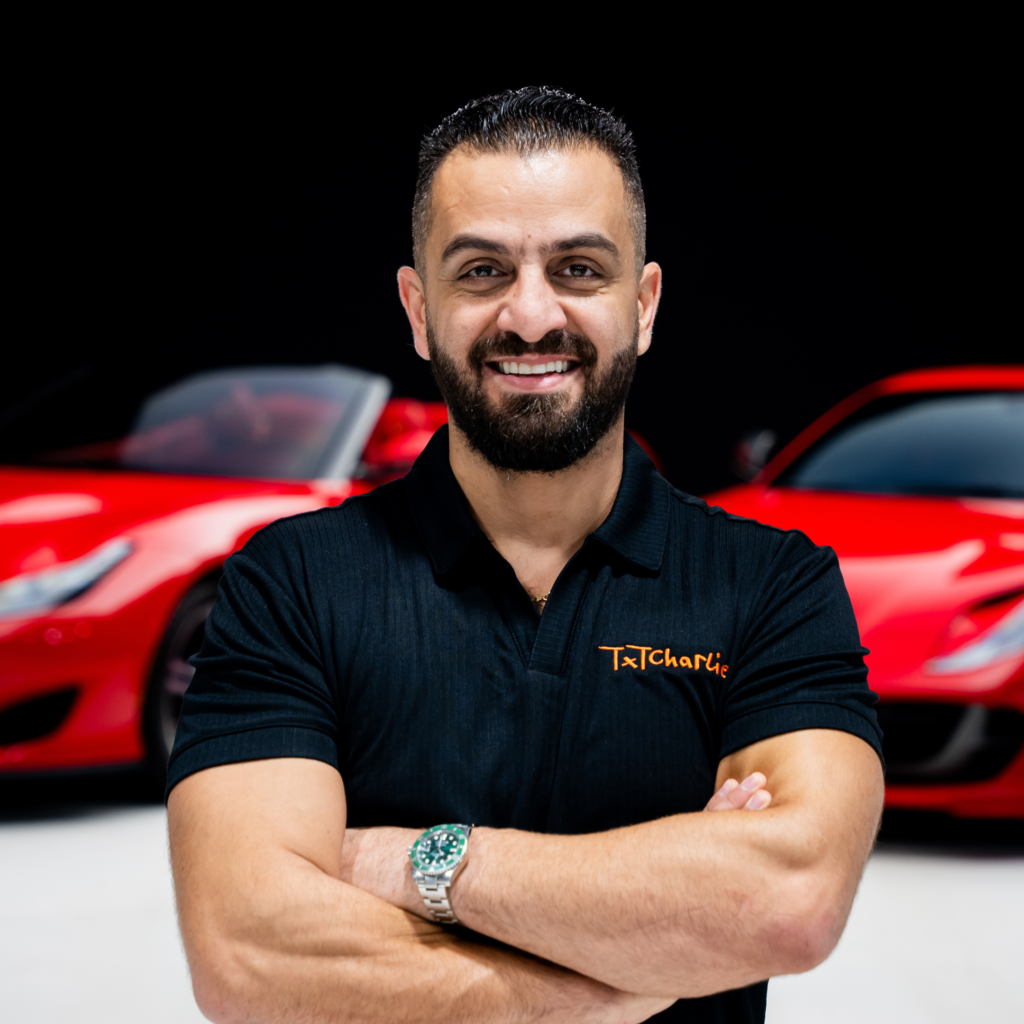20png | Txtcharlie Smiling man with dark hair and beard, arms crossed, wearing a black polo; red cars blurred in the background.