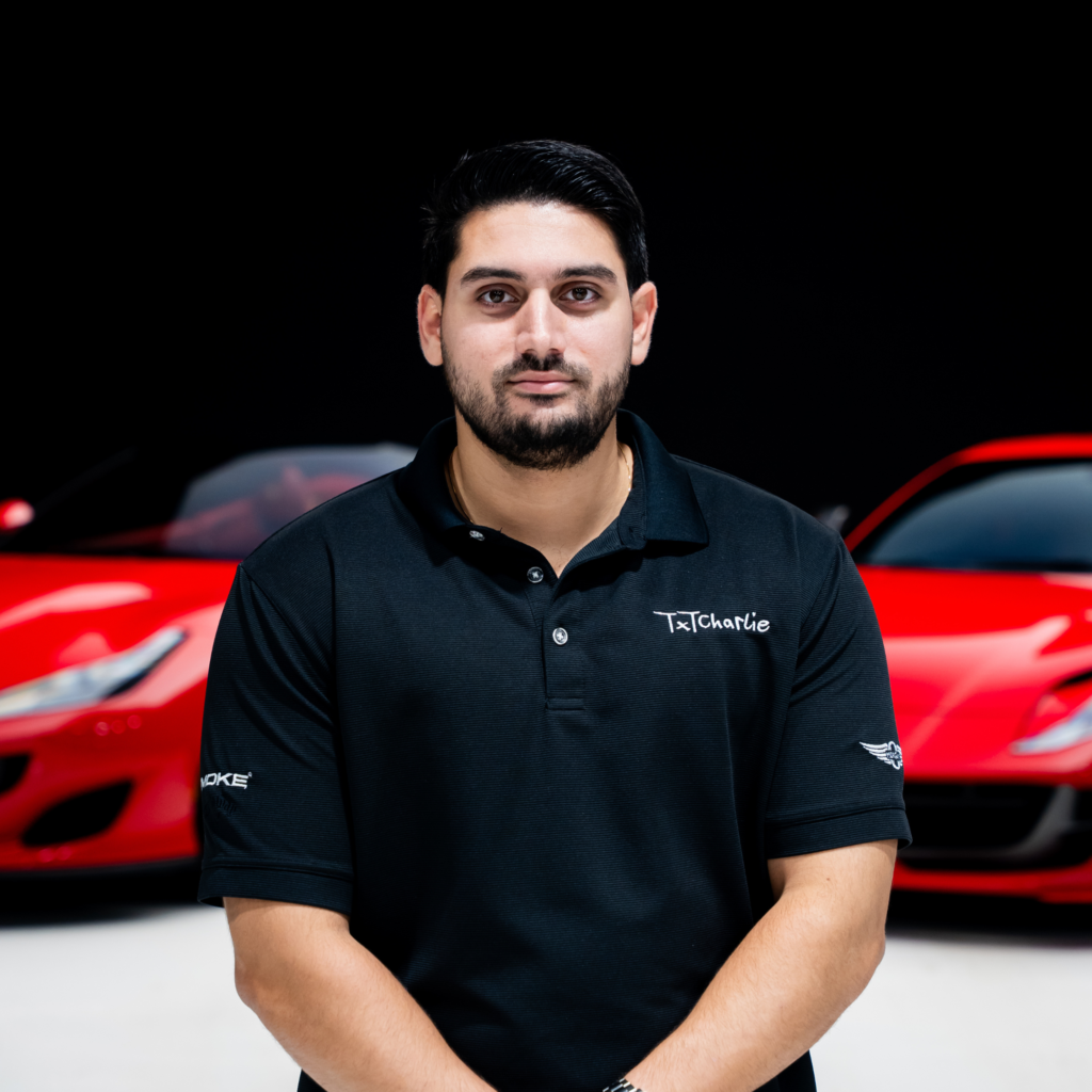 22png | Txtcharlie Portrait of a man with short black hair and a beard, wearing a black polo shirt, standing in front of a red car.