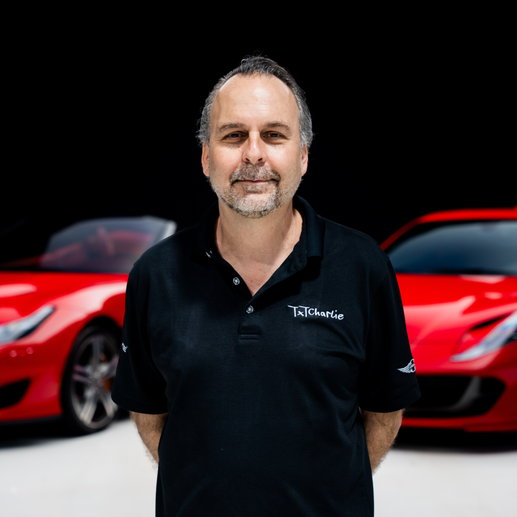 231png | Txtcharlie Smiling man with gray beard in a black polo shirt, standing in front of red cars in a showroom background.