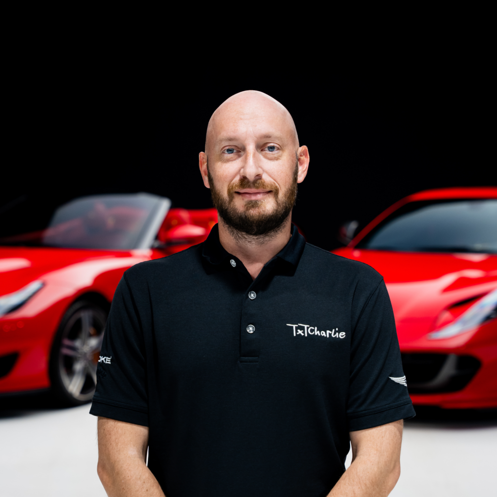 262png | Txtcharlie Portrait of a bald man with a beard in a black TXCharlie polo, standing in front of red sports cars in a showroom.