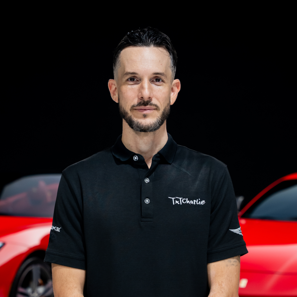 271png | Txtcharlie Man with dark hair and a beard wearing a black polo, standing in front of a red car on display.