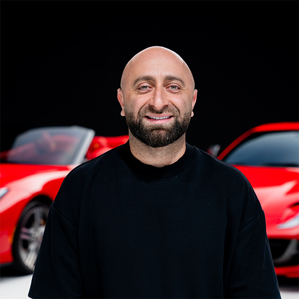 3png | Txtcharlie Portrait of a smiling bald man with a beard wearing a black shirt, standing in front of red sports cars behind him.