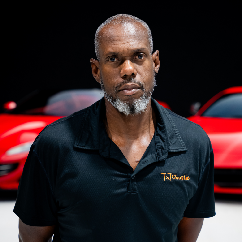 6png | Txtcharlie Close-up of a man in a black polo with orange 'TxCharlie' embroidery on the chest, in front of a red car.