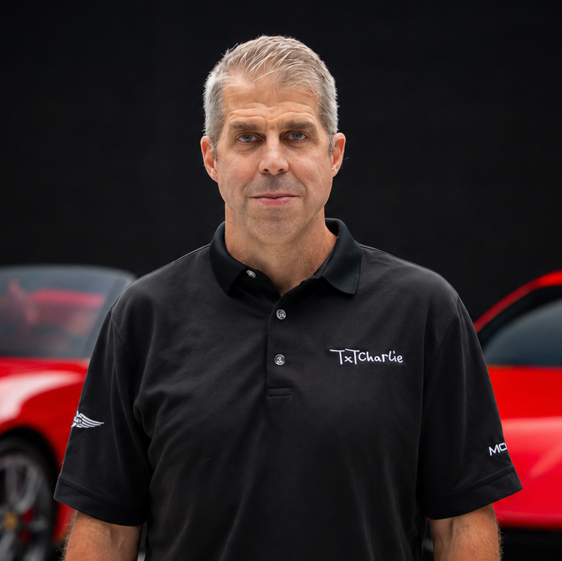 7da15591bee242a196df0ecfddab559bpng | Txtcharlie Man with short gray hair wearing a black polo shirt with a logo, standing in front of a red car behind him.