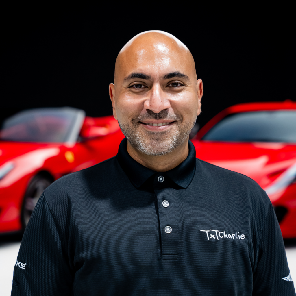 8png | Txtcharlie Close-up portrait of a smiling man with a bald head wearing a black polo, 'TxCharlie' visible on the chest, with a red car blurred in the background.