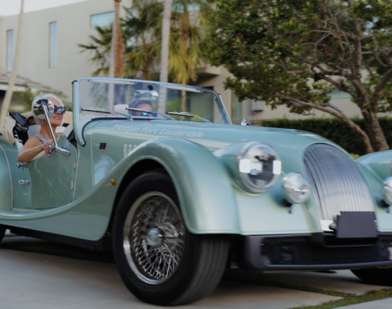 Two people ride in a vintage teal convertible driving down a suburban street; driver wears sunglasses and smiles, passenger in the passenger seat also visible.