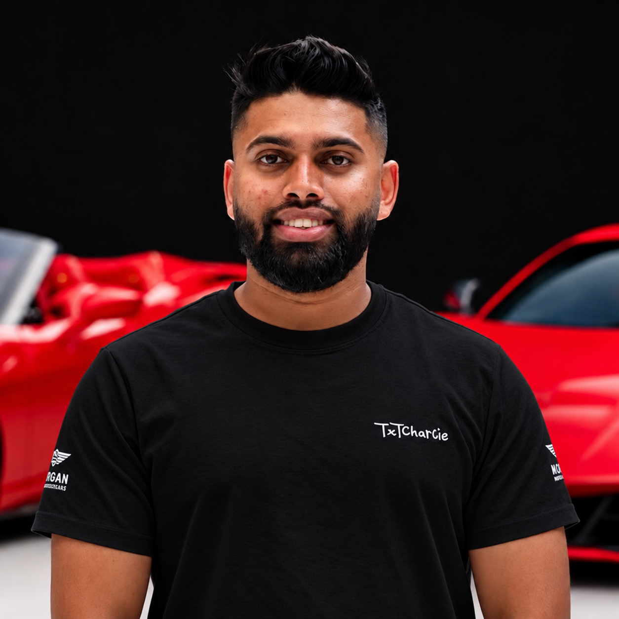 BhudeshShilchand02 Portrait of a smiling man with a beard and styled hair, wearing a black T-shirt with a white logo, standing in front of red cars in a showroom.