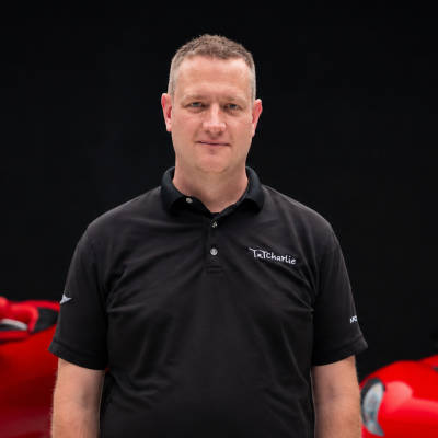 Portrait of a man in a black polo shirt with a TexCharlie logo, standing against a dark background with red cars blurred in the background.