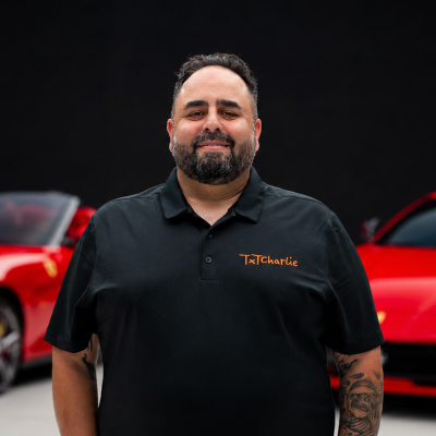 Smiling man with a beard in a black polo with an orange 'TexCharlie' logo, standing in front of red cars.