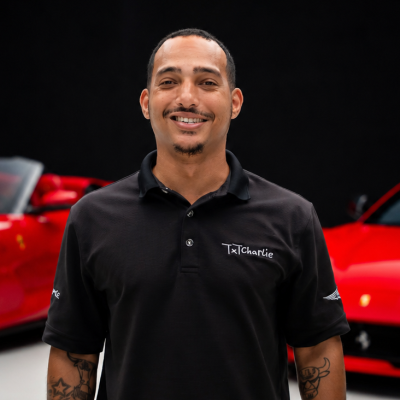 Portrait of a smiling man in a black polo shirt with a TxCharlie logo, standing in front of red cars in a showroom.