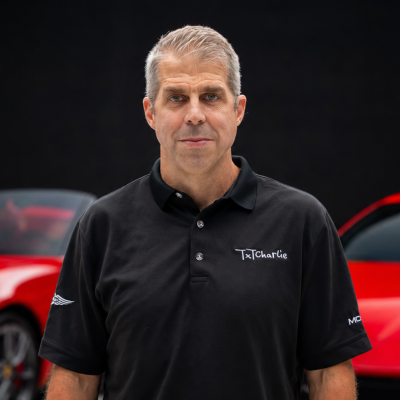 Man with short gray hair wearing a black polo in front of a red sports car; shirt reads 'TexCharlie'.|Portrait of a middle-aged man in a black polo, standing outdoors with a red car in the background.